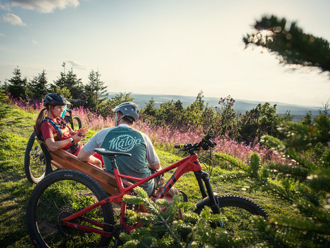 Stoneman_Miriquidi_c_TMGS_Philipp Ruopp_2020 (27).jpg Zwei Mountainbiker pausieren auf einer Bank auf einem bewaldeten Hügel mit Fernsicht.