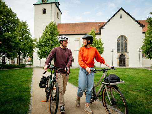 Zwei Radfahrer mit Helmen gehen entlang eines Pfades vor einer historischen Kirche.