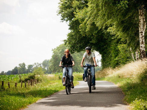 Zwei Männer fahren lachend auf einer asphaltierten Landstraße durch grüne Landschaft.