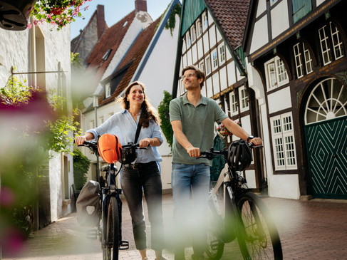 Zwei Personen laufen mit Fahrrädern durch eine gepflasterte Straße mit Fachwerkhäusern.Two people walk with bicycles through a cobbled street with half-timbered houses.To personer går med cykler gennem en brostensbelagt gade med bindingsværkshuse.Twee mensen lopen met fietsen door een geplaveide straat met vakwerkhuizen.