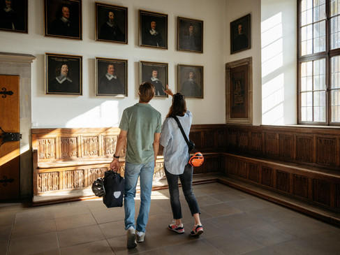 Zwei Besucher betrachten Gemälde in einem historischen Raum mit Holztäfelung und großen Fenstern.