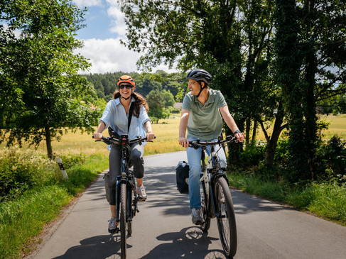 Zwei Radfahrer auf einer ländlichen Straße, umgeben von grünen Bäumen und Feldern.