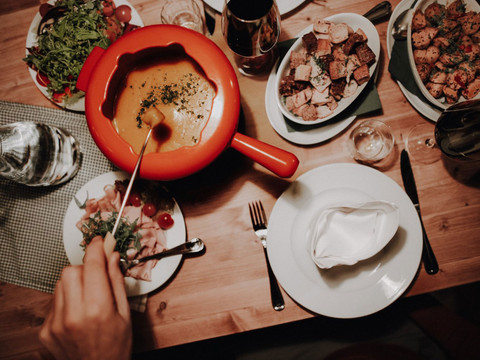 Ein rustikales Fondue auf Holztisch, umgeben von Beilagen, Kräutern und Rotwein in gemütlicher Atmosphäre.A rustic fondue on a wooden table, surrounded by side dishes, herbs and red wine in a cozy atmosphere.Rustikální fondue na dřevěném stole, obklopené přílohami, bylinkami a červeným vínem v útulné atmosféře.Rustykalne fondue na drewnianym stole, otoczone dodatkami, ziołami i czerwonym winem w przytulnej atmosferze.Een rustieke fondue op een houten tafel, omringd door bijgerechten, kruiden en rode wijn in een gezellige sfeer.Una fonduta rustica su un tavolo di legno, circondata da contorni, erbe e vino rosso in un'atmosfera accogliente.