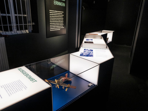 Museumsausstellung mit beleuchteten Glasvitrinen, die Texte und ein Modellflugzeug präsentieren.