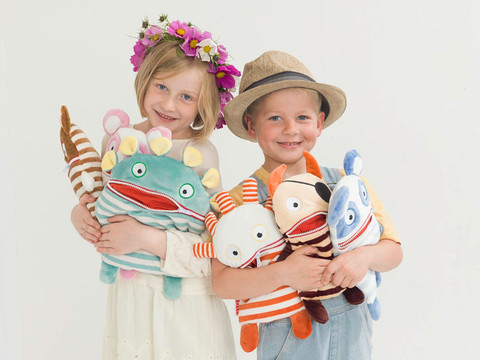 Sorgenfresser Zwei lächelnde Kinder mit Blumenkranz und Hut halten bunte Plüschfiguren vor neutralem Hintergrund.