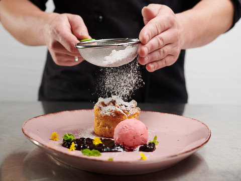Parkhotel Neustadt Dessert Ein Koch bestreut ein köstliches Dessert mit Puderzucker, begleitet von Sorbet und Beeren auf einem Teller.
