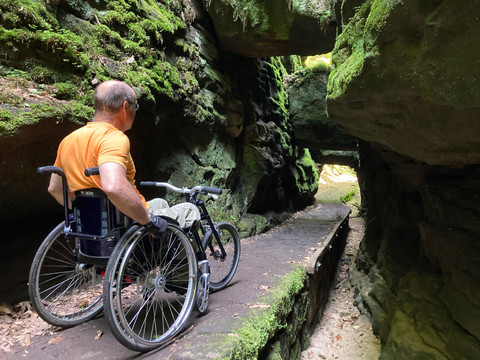 Skalní brána Ein Rollstuhlfahrer steht auf einem Wanderweg unter einem natürlichen Tor aus Sandsteinblöcken. A wheelchair user stands on a hiking trail under a natural gate made of sandstone blocks.Vozíčkář stojí na chodníku pod přírodní bránou z pískovcových kvádrů.Een rolstoelgebruiker staat op een voetpad onder een natuurlijke poort van zandsteenblokken.