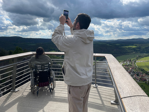 Rollstuhl Ein Mann fotografiert die Landschaft, ein Rollstuhlfahrer blickt von der Aussicht in die Landschaft. A man photographs the landscape, a wheelchair user looks out from the view.Muž fotografuje krajinu, z výhledu se dívá vozíčkář.Een man fotografeert het landschap, een rolstoelgebruiker kijkt uit op het uitzicht.