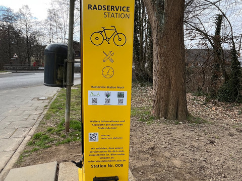 Radservice-Station Die Radservice-Station ist eine gelbe, aufrecht stehende Servicestation mit ADAC-Logo und Manometer. Im Vordergrund sind Fahrradwerkzeuge angebracht, während dahinter ein asphaltierter Gehweg und Bäume zu sehen sind.