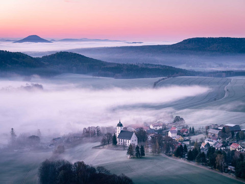 Uitzicht op Papstdorf vanaf de Papststein Nebelbedeckte Landschaft bei Sonnenaufgang mit Kirche und Papstdorf im Vordergrund, Hügel im Hintergrund.