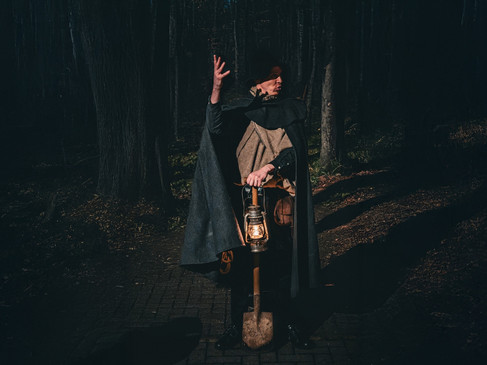 Ein Mann in historischer Kleidung mit Umhang und Laterne steht in einem dunklen Wald auf einem Weg.