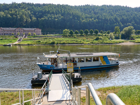 Fähre am Ufer der Elbe, mit bewaldeten Hügeln im Hintergrund und einem historischen Gebäude am Fluss.