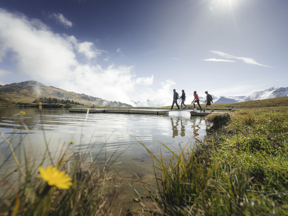Randonnée Foxtrail au bord du lac Foxtrail Desoria Aletsch Arena mit vier Personen, die über einen Steg am Bettmersee wandern, vor den Walliser AlpenFoxtrail Desoria Aletsch Arena with four people hiking over a footbridge on Lake Bettmersee, in front of the Valais AlpsFoxtrail Desoria Aletsch Arena avec quatre personnes marchant sur une passerelle au Bettmersee, devant les Alpes valaisannes