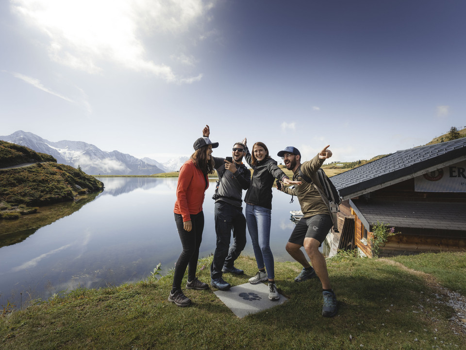 Foxtrail group at Bettmersee Foxtrail Desoria Aletsch Arena mit vier Teilnehmern am Bettmersee, lachend vor der Kulisse der Walliser AlpenFoxtrail Desoria Aletsch Arena with four participants at Lake Bettmersee, laughing against the backdrop of the Valais AlpsFoxtrail Desoria Aletsch Arena avec quatre participants au Bettmersee, riant devant le décor des Alpes valaisannes