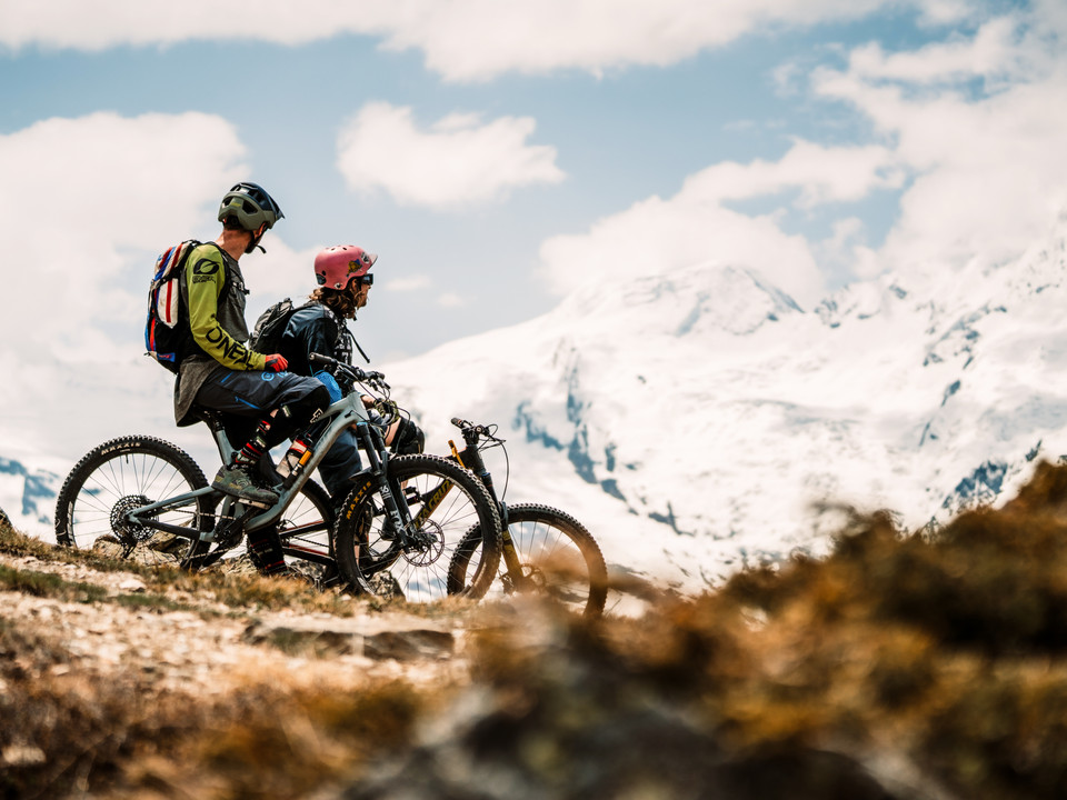 Biken vor verschneiter Alpenkulisse Oberwalliser Bikepass mit Bikern auf alpinem Trail mit Blick auf schneebedeckte Gipfel der Walliser Alpen in Saas FeeUpper Valais bike pass with bikers on an alpine trail with views of the snow-capped peaks of the Valais Alps in Saas FeeBikepass haut-valaisan avec des vététistes sur un trail alpin avec vue sur les sommets enneigés des Alpes valaisannes à Saas Fee
