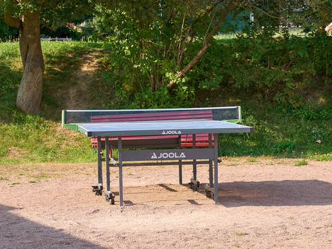 Tischtennisplatte im Park auf sandigem Boden, umgeben von Gras und Bäumen.