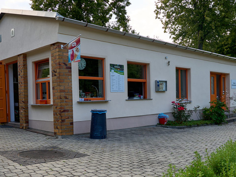 Imbiss und Eingang Stadtbad Hohnstein Kleines weißes Gebäude mit orangefarbenen Türen und Fensterrahmen, umgeben von Grünanlagen.