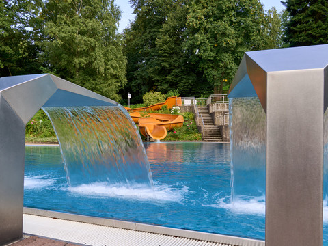 Zwei Wasserfontänen fließen in einen Pool, im Hintergrund eine orange Rutsche.