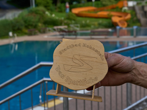 Stadtbad Hohnstein Hand hält hölzernes Emblem mit Aufschrift "Freibad Hohnstein -das Sportliche" vor Schwimmbad.