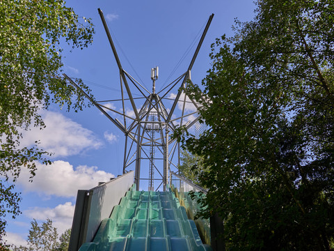Metallturm mit grünen Rutschen zwischen Bäumen unter blauem Himmel.