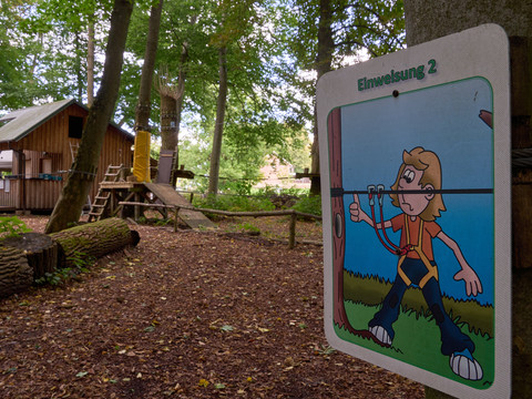 Schild mit Balancierübung im Kletterwald, Holzhütte im Hintergrund.