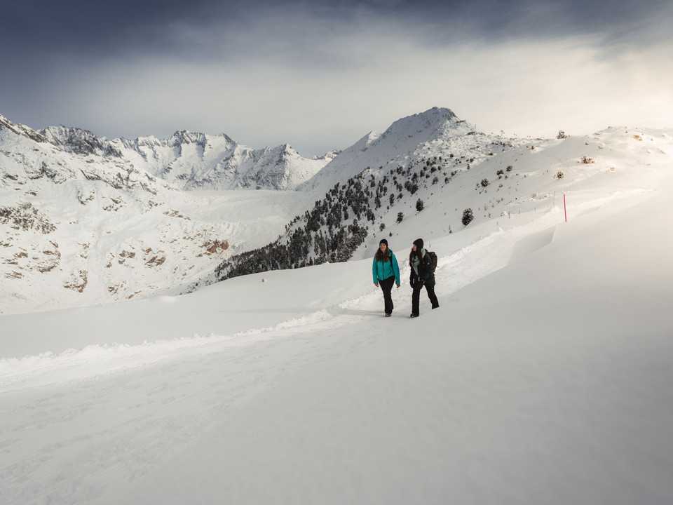 Winterwandern mit Aussicht Aletsch Entdeckerpass mit zwei Frauen beim Winterwandern in der verschneiten Landschaft mit Blick auf den Aletschgletscher