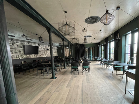 Taverne.jpg Ruime eetzaal met houten vloer, veel tafels en stoelen, ramen van vloer tot plafond en groene pilaren in het midden.