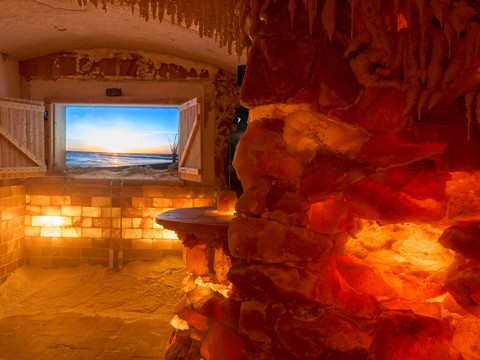 Salzgrotte Lindenhof Königstein Innenraum einer Salzgrotte mit beleuchteten Salzsteinwänden und TV-Fensterblick auf den Sonnenuntergang am Meer.Interior of a salt cave with illuminated salt stone walls and TV window view of the sunset by the sea.Interiér solné jeskyně s osvětlenými stěnami ze solných kamenů a výhledem z okna televize na západ slunce u moře.Wnętrze jaskini solnej z podświetlonymi ścianami z kamienia solnego i widokiem z okna telewizora na zachód słońca nad morzem.Interieur van een zoutgrot met verlichte zoutstenen muren en tv-raam met uitzicht op de zonsondergang aan zee.Interno di una grotta di sale con pareti di pietra salina illuminate e finestra TV con vista sul tramonto in riva al mare.