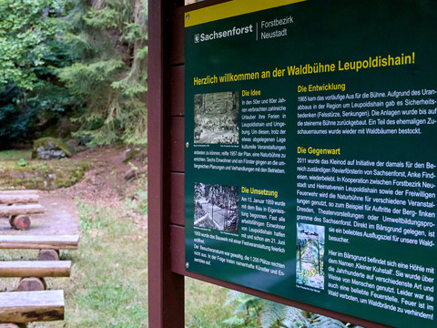 Waldbühne Leupoldishain Informationsstele im Vordergrund, Holzbänke und Bäume im Hintergrund.