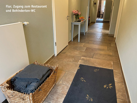 Gang.jpg breiter Flur mit rotbraunen Fliesen, einem schwarzen Läufer, einem Korb voller Decken, einem Tischchen mit Blumengesteck sowie mehreren weißen TürenWide hallway with red-brown tiles, a black runner, a basket full of blankets, a small table with a flower arrangement and several white doorsŠiroká chodba s červenohnědou dlažbou, černým běhounem, košem plným dek, malým stolkem s květinovou výzdobou a několika bílými dveřmi.Brede hal met roodbruine tegels, een zwarte loper, een mand vol dekens, een kleine tafel met een bloemstuk en verschillende witte deuren