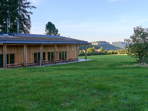 Walderlebniszentrum Holzgebäude mit großen Fenstern neben grüner Wiese und Bäumen.Wooden building with large windows next to a green meadow and trees.Dřevěná budova s velkými okny vedle zelené louky a stromů.Drewniany budynek z dużymi oknami obok zielonej łąki i drzew.Houten gebouw met grote ramen naast een groene weide en bomen.Edificio in legno con grandi finestre accanto a un prato verde e ad alberi.