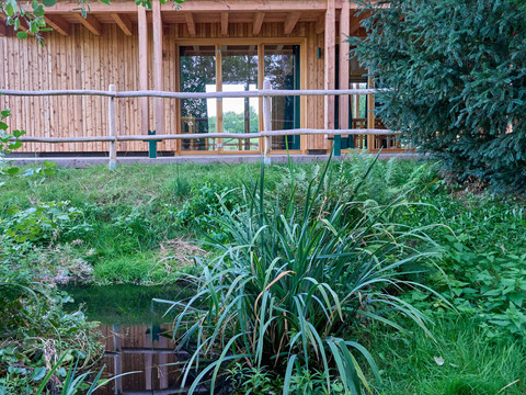 Walderlebniszentrum Holzhaus mit großen Fenstern umgeben von Grünflächen und einem kleinen Teich.Wooden house with large windows surrounded by green areas and a small pond.Dřevěný dům s velkými okny obklopený zelení a malým rybníkem.Drewniany dom z dużymi oknami otoczony terenami zielonymi i małym stawem.Houten huis met grote ramen omgeven door groene zones en een kleine vijver.Casa in legno con ampie finestre circondata da aree verdi e da un piccolo stagno.