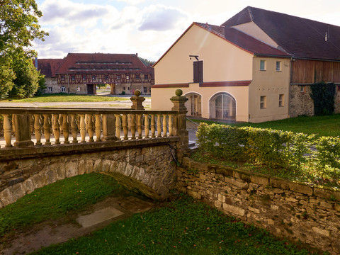 Kulturscheune und Schloss Langburkersdorf Steinbrücke mit Geländer vor historischen Gebäuden und grüner Wiese.Stone bridge with railings in front of historic buildings and a green meadow.Kamenný most se zábradlím před historickými budovami a zelenou loukou.Kamienny most z balustradami przed zabytkowymi budynkami i zieloną łąką.Stenen brug met hekwerk voor historische gebouwen en een groene weide.Ponte in pietra con ringhiera davanti a edifici storici e a un prato verde.
