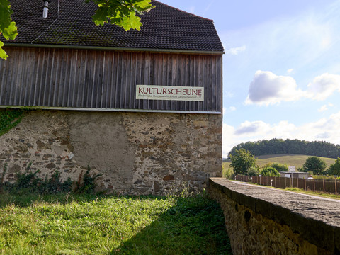 Steinmauer im Vordergrund, dahinter Scheune mit Schild "KULTURSCHEUNE", Landschaft im Hintergrund.Stone wall in the foreground, behind it barn with sign "KULTURSCHEUNE", landscape in the background.V popředí kamenná zeď, za ní stodola s nápisem "KULTURSCHEUNE", v pozadí krajina.Kamienna ściana na pierwszym planie, za nią stodoła z napisem "KULTURSCHEUNE", w tle krajobraz.Stenen muur op de voorgrond, daarachter schuur met bord "KULTURSCHEUNE", landschap op de achtergrond.Muro di pietra in primo piano, dietro di esso fienile con insegna "KULTURSCHEUNE", paesaggio sullo sfondo.