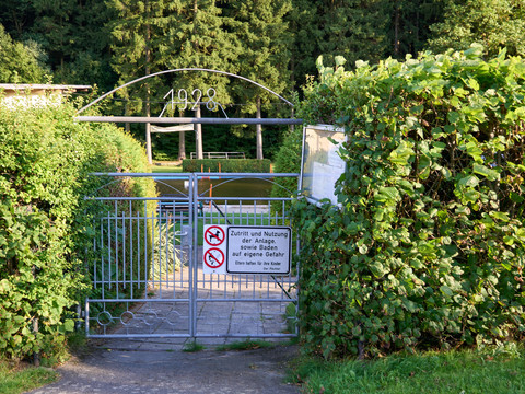 Waldbad Eingang Verschlossenes Tor mit Schild "Kein Zutritt für Unbefugte" umgeben von grünen Hecken.Locked gate with sign "No access for unauthorized persons" surrounded by green hedges.Uzamčená brána s nápisem "Nepovolaným osobám vstup zakázán" obklopená zeleným živým plotem.Zamknięta brama z napisem "Nieupoważnionym wstęp wzbroniony" otoczona zielonym żywopłotem.Afgesloten hek met bordje "Geen toegang voor onbevoegden" omgeven door groene heggen.Cancello chiuso con cartello "Vietato l'accesso ai non autorizzati", circondato da siepi verdi.