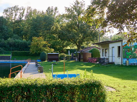 Waldbad Freibad mit grünem Rasen und Graffiti an einer Wand, umgeben von Bäumen.Outdoor pool with green lawn and graffiti on a wall, surrounded by trees.Venkovní bazén se zeleným trávníkem a graffiti na zdi, obklopený stromy.Odkryty basen z zielonym trawnikiem i graffiti na ścianie, otoczony drzewami.Buitenzwembad met groen gazon en graffiti op een muur, omringd door bomen.Piscina all'aperto con prato verde e graffiti su un muro, circondata da alberi.