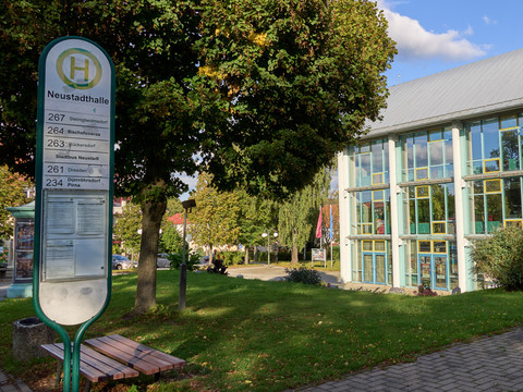 Neustadthalle Bushaltestelle mit Fahrplan vor einem Gebäude mit großer Glasfassade und Bäumen.