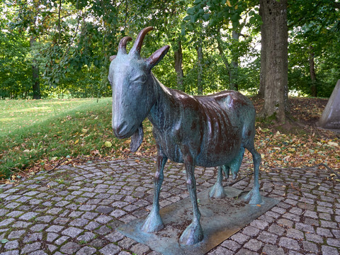 Bronzestatue Ziege auf einem gepflasterten Weg mit Bäumen im Hintergrund.