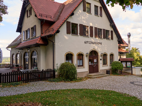 Weißes Gebäude mit braunem Dach und "Götzinger Höhe" Schriftzug, umgeben von Bäumen.