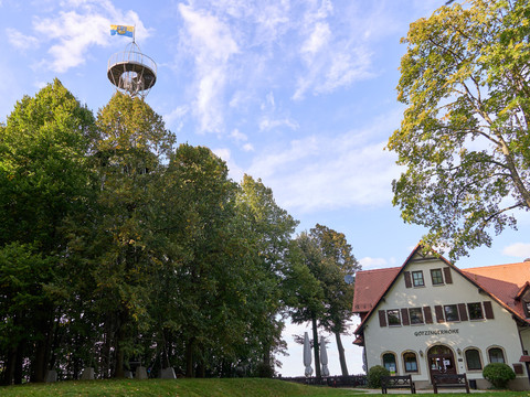 Aussichtsturm hinter Bäumen neben einem Haus mit Schild "Götzinger Höhe".