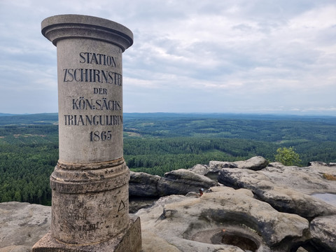 Steinsäule mit Inschrift "Station Zschirnstein der Kön. Sächs. Triangulirung 1865" vor Aussicht auf Waldlandschaft.