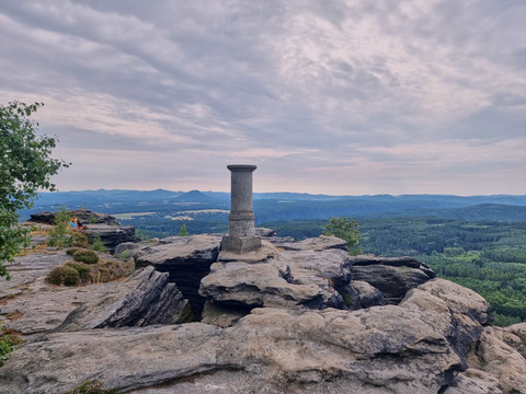 Großer Zschirnstein Aussicht Steinerner Säulenstumpf auf Felsvorsprung mit bewölktem Himmel und Waldlandschaft im Hintergrund.
