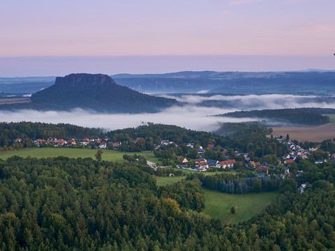 Blick vom Gohrisch nach Kurort Gohrisch Landschaft mit Nebel über Gohrisch, umgeben von Wäldern und einem Berg im Hintergrund bei Sonnenaufgang.