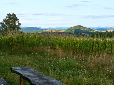 Holzbank im Vordergrund mit Blick auf grüne Hügel und bewölkten Himmel.Wooden bench in the foreground with a view of green hills and a cloudy sky.Dřevěná lavička v popředí s výhledem na zelené kopce a zataženou oblohu.Drewniana ławka na pierwszym planie z widokiem na zielone wzgórza i zachmurzone niebo.Houten bank op de voorgrond met uitzicht op groene heuvels en een bewolkte lucht.Panchina di legno in primo piano con vista su colline verdi e cielo nuvoloso.