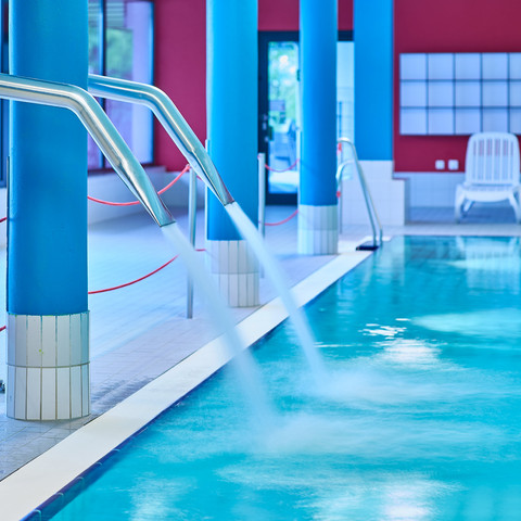 Nichtschwimmerbecken Oktopus Das Bild zeigt den Innenbereich eines Schwimmbads mit leuchtend blauen Säulen und einem ruhigen Wasserbecken. Im Hintergrund sind zwei weiße Liegestühle vor einer roten Wand sichtbar.