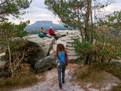Frau wandert auf Pfad zu zwei Personen, die auf einem Felsen in bergiger Landschaft sitzen.