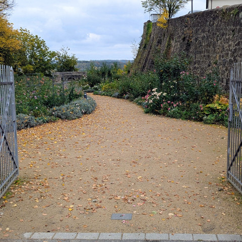 ©Bastian Ulrich - Rosengarten 2 Offener schmiedeeiserner Torbogen, der einen kiesbedeckten Pfad zwischen alten Steinmauern freigibt. Der Weg ist gesäumt von bunten Herbstblättern und üppiger Vegetation.