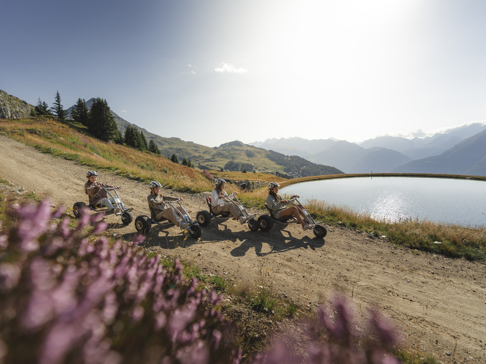 Mountaincart-Tour am Speichersee auf der Riederalp Vier Freunde fahren entlang des glitzernden Speichersees mit dem Mountaincart Aletsch Arena auf der Riederalp, umgeben von Alpenblumen und SommerlichtFour friends ride along the glistening reservoir on the Aletsch Arena mountain cart on the Riederalp, surrounded by alpine flowers and summer lightQuatre amis longent le lac de barrage scintillant en mountaincart Aletsch Arena à Riederalp, entourés de fleurs alpines et de lumière d'été.