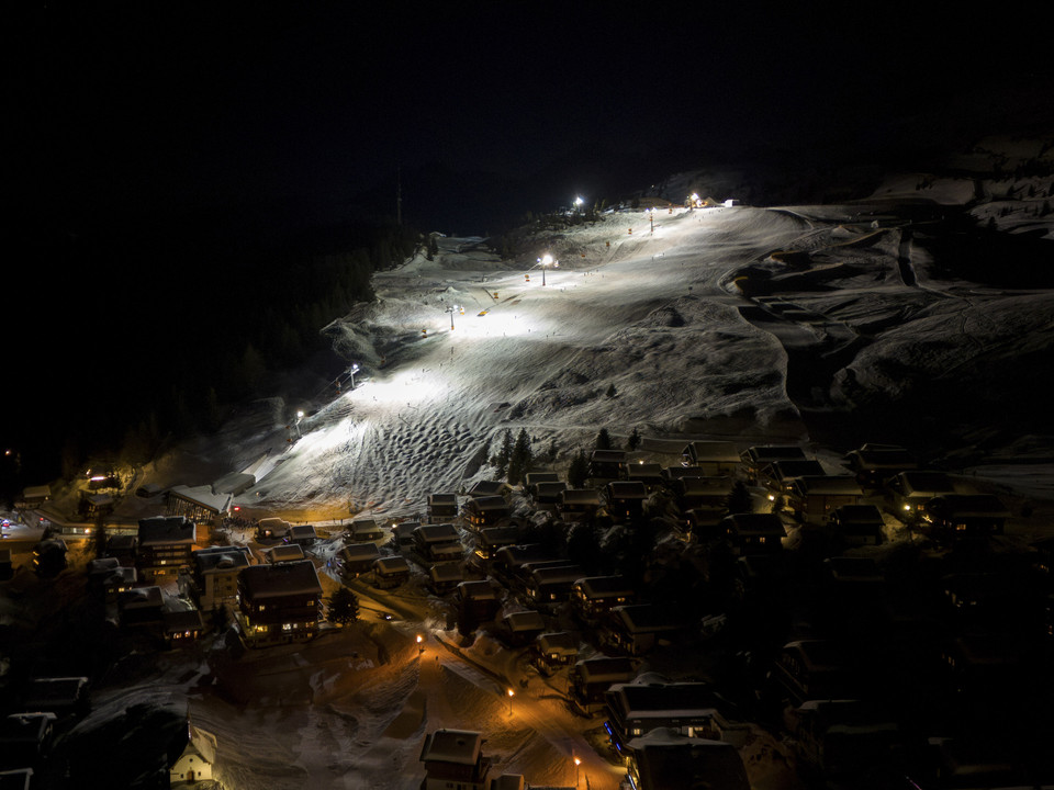 Illuminated ski slopes for night skiing on the Bettmeralp Das Nachtskifahren Bettmeralp Riederalp zeigt von oben die hell erleuchteten Skipisten und Chalets der Aletsch Arena an einem klaren WinterabendNight skiing Bettmeralp Riederalp shows the brightly lit ski slopes and chalets of the Aletsch Arena from above on a clear winter eveningLe ski nocturne Bettmeralp Riederalp montre d'en haut les pistes de ski et les chalets de l'Aletsch Arena éclairés par une claire soirée d'hiver.