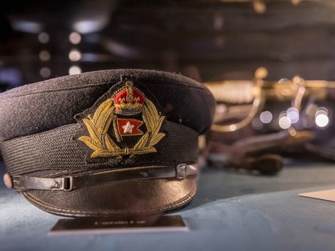 Eine Kapitänsmütze mit goldener Verzierung und rotem Abzeichen der Titanic in einer Ausstellungsvitrine.A captain's cap with golden decorations and a red Titanic badge in a display case.Kapitánská čepice se zlatými ozdobami a červeným odznakem Titanic ve vitríně.Czapka kapitańska ze złotymi zdobieniami i czerwoną odznaką Titanica w gablocie.Een kapiteinspet met gouden versieringen en een rode Titanic-badge in een vitrinekast.Un berretto da capitano con decorazioni dorate e un distintivo rosso del Titanic in una vetrina.